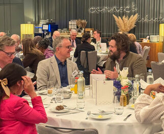 Eine Gruppe von Menschen sitzt an einem Tisch in einem eleganten Restaurant. Einige Personen unterhalten sich, während andere essen. Der Tisch ist mit Geschirr, Gläsern und Blumen dekoriert. Im Hintergrund sind weitere Gäste und eine stilvolle Einrichtung zu sehen.