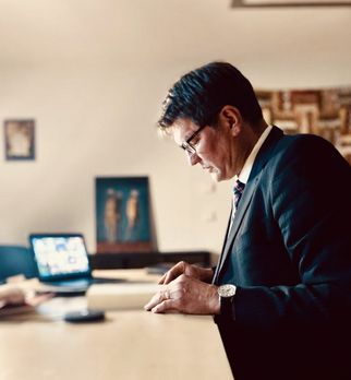 Ein Mann in einem Anzug steht an einem Tisch und schaut auf ein Buch. Im Hintergrund ist ein Laptop und einige Bilder an der Wand zu sehen. Der Mann trägt eine Brille und hat eine nachdenkliche Haltung. (Text KI generiert)