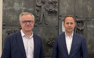 Zwei Männer stehen nebeneinander vor einer Wand mit reliefartigen Metallplatten, die verschiedene Szenen darstellen. Beide tragen Anzüge und schauen in die Kamera. (Text KI generiert)