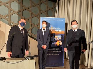 Drei Männer in Anzügen und Mund-Nasen-Schutz stehen vor einem Banner der Initiative 27. Januar. Der Banner thematisiert das Gedenken an den Holocaust und den Kampf gegen Antisemitismus. Die Männer posieren in einem Veranstaltungsraum mit Mikrofonen und einem Podium im Hintergrund. (Text KI generiert)