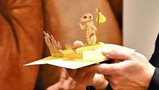 Eine Frau in schwarzer Kleidung gestikuliert, während ein Mann in einem Anzug ein aufklappbares Kunstwerk mit einem Tier hält. Im Hintergrund ist ein Gemälde zu sehen. Beide Personen scheinen sich über das Kunstwerk zu freuen. (Text KI generiert)