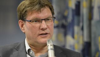 Ein Mann mit Brille und Anzug spricht in ein Mikrofon. Er hat kurzes, braunes Haar und gestikuliert während des Gesprächs. Im Hintergrund sind unscharfe Vorhänge zu sehen. (Text KI generiert)