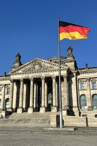 Das Bild zeigt das Reichstagsgebäude in Berlin mit einer großen deutschen Flagge im Vordergrund. Der Himmel ist klar und blau, und die Architektur des Gebäudes ist im klassischen Stil gehalten, mit Säulen und einem markanten Dach.