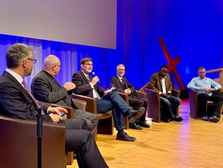 Eine Diskussionsrunde mit mehreren Männern, die auf Stühlen sitzen. Einer der Männer spricht ins Mikrofon, während die anderen aufmerksam zuhören. Im Hintergrund ist eine große Leinwand und eine rote Kreuzskulptur zu sehen. Die Atmosphäre ist formell und konzentriert. (Text KI generiert)