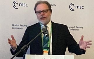Ein Mann steht an einem Rednerpult mit der Aufschrift "International Prayer Breakfast" und spricht. Er trägt einen Anzug und eine Krawatte, hat eine Brille auf und gestikuliert mit den Händen. Im Hintergrund sind Logos der Münchner Sicherheitskonferenz zu sehen. (Text KI generiert)