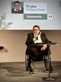 Ein älterer Mann sitzt im Rollstuhl und spricht in ein Mikrofon. Er trägt eine Brille und einen schwarzen Anzug. Im Hintergrund ist eine Präsentation zu sehen, die seinen Namen und das Jubiläum „75 Jahre“ zeigt. (Text KI generiert)