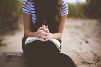 Eine Person sitzt im Sand und hält ein aufgeschlagenes Buch auf ihrem Schoß. Die Hände sind gefaltet und die Person trägt ein gestreiftes T-Shirt. Im Hintergrund sind unscharfe Gräser zu sehen. (Text KI generiert)