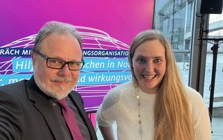 Zwei Personen posieren für ein Selfie vor einem großen Bildschirm mit einem pinken Hintergrund. Auf dem Bildschirm steht ein Text über Unterstützung und Organisation. Die Person links trägt einen Anzug und eine Krawatte, die Person rechts hat lange, blonde Haare und trägt einen weißen Pullover. (Text KI generiert)