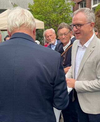 Eine Gruppe von Menschen steht im Freien, einige sprechen miteinander. Ein Mann in einem hellen Anzug lächelt und hält ein Dokument in der Hand. Im Hintergrund sind weitere Personen und ein Zelt zu sehen. Die Atmosphäre wirkt freundlich und gesellig. (Text KI generiert)