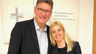 Zwei Personen stehen lächelnd nebeneinander in einem Büro. Der Mann trägt einen Anzug und hat eine Brille, die Frau ist in schwarzer Kleidung gekleidet. Im Hintergrund ist ein Schild mit dem Text "Die Evangelische Allianz in Deutschland" zu sehen. (Text KI generiert)