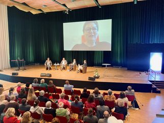 Eine Veranstaltung in einem großen Saal mit Publikum. Auf der Bühne sitzen vier Personen, während eine weitere Person über einen Bildschirm zugeschaltet ist. Der Raum ist gut beleuchtet und es gibt Pflanzen und Tische auf der Bühne. (Text KI generiert)