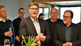 Eine Gruppe von fünf Personen steht in einem Raum. Ein Mann in einem Anzug spricht und gestikuliert, während die anderen aufmerksam zuhören. Auf dem Tisch vor ihnen stehen Getränke und eine Pflanze. Die Atmosphäre wirkt gesellig und informell. (Text KI generiert)