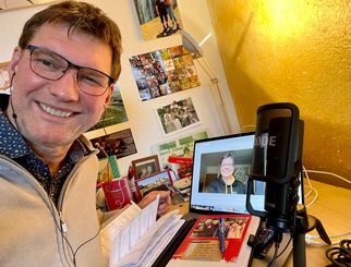 Ein lächelnder Mann mit Brille sitzt an einem Schreibtisch. Er hält Unterlagen in der Hand und schaut auf einen Laptop, auf dem ein Bild einer anderen Person zu sehen ist. Neben dem Laptop steht ein Mikrofon. Im Hintergrund sind Fotos an der Wand. (Text KI generiert)