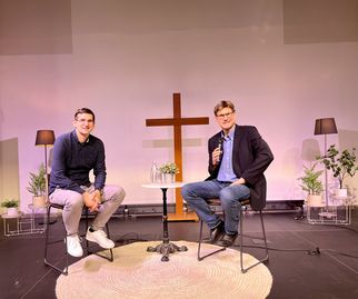 Zwei Männer sitzen auf Stühlen auf einer Bühne. Im Hintergrund steht ein Kreuz. Der Mann links trägt ein kariertes Hemd und lächelt, während der Mann rechts ein Mikrofon hält und in einem Anzug sitzt. Pflanzen und Lampen sind um sie herum angeordnet. (Text KI generiert)