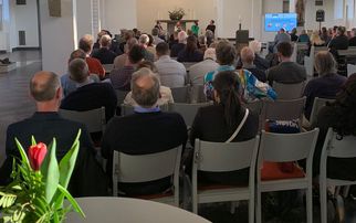 Eine Gruppe von Menschen sitzt in einem hellen Raum mit hohen Fenstern und einer hohen Decke. Im Vordergrund steht ein Tisch mit einer Blumendekoration. Im Hintergrund ist eine Präsentation auf einem Bildschirm zu sehen, während eine Person spricht. (Text KI generiert)