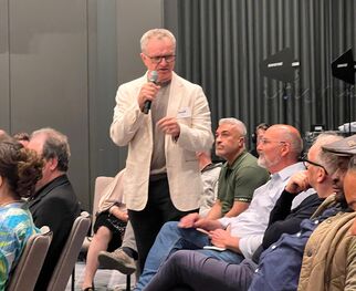 Eine Gruppe von Menschen sitzt in einem Konferenzraum. Ein Mann mit Brille und einem Mikrofon spricht, während andere Zuhörer aufmerksam zuhören. Die Atmosphäre wirkt informell und interaktiv.