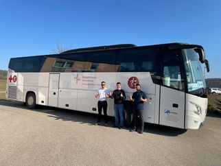 Drei Personen stehen vor einem großen, weißen Reisebus mit einem roten Kreuz-Symbol. Der Bus ist auf einem Parkplatz geparkt, und die Personen zeigen Daumen hoch. Der Himmel ist klar und blau. (Text KI generiert)