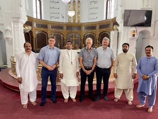 Eine Gruppe von sechs Männern steht in einem religiösen Gebäude. Sie tragen traditionelle Kleidung und stehen vor einem kunstvoll gestalteten Altar mit religiösen Symbolen. Der Raum hat hohe Fenster und eine festliche Atmosphäre. (Text KI generiert)