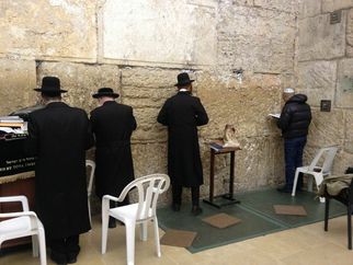 Vier Männer in traditionellen schwarzen Anzügen und Hüten beten an einer Steinmauer. Im Hintergrund steht ein Tisch mit religiösen Texten. Ein weiterer Mann in blauer Kleidung sitzt auf einem Stuhl und liest. Der Raum hat einen schlichten, historischen Charakter. (Text KI generiert)