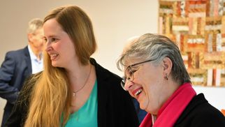 Zwei Frauen stehen in einem hellen Raum und lächeln. Die Frau links hat lange, blonde Haare und trägt ein grünes Kleid. Die Frau rechts trägt eine schwarze Jacke und einen pinken Schal. Im Hintergrund sind unscharfe Personen und eine dekorative Wandgestaltung zu sehen. (Text KI generiert)