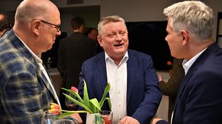 Drei Männer stehen an einem Tisch und unterhalten sich. Auf dem Tisch stehen Gläser, Snacks und eine Vase mit Tulpen. Die Männer tragen formelle Kleidung und scheinen in ein angeregtes Gespräch vertieft zu sein. (Text KI generiert)