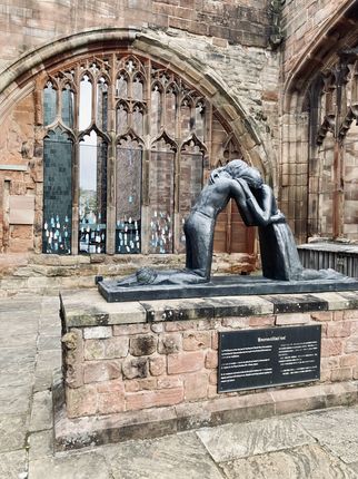 Eine bronzene Skulptur zeigt zwei sich umarmende Figuren in einer emotionalen Geste. Im Hintergrund sind gotische Fenster und alte Steinmauern sichtbar. Die Szene vermittelt ein Gefühl von Versöhnung und Nähe. (Text KI generiert)