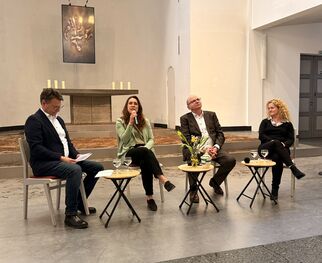Eine Diskussionsrunde mit vier Personen in einem hellen Raum. Zwei Männer und zwei Frauen sitzen an kleinen Tischen. Eine Frau spricht, während die anderen zuhören. Im Hintergrund sind Kerzen und ein Kunstwerk an der Wand zu sehen. (Text KI generiert)