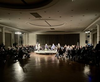 Eine Gruppe von Menschen sitzt in einem großen Raum, um einen runden Tisch herum. Auf der Bühne sind mehrere Personen, die diskutieren. Der Raum ist schwach beleuchtet, mit Scheinwerfern, die auf die Bühne gerichtet sind. Zuschauer sitzen in Stuhlreihen und hören aufmerksam zu. (Text KI generiert)