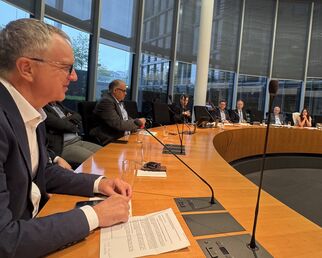 Eine Gruppe von Personen sitzt an einem großen, runden Tisch in einem Konferenzraum. Im Vordergrund ist ein Mann zu sehen, der spricht. Im Hintergrund sind weitere Teilnehmer, die aufmerksam zuhören. Große Fenster lassen Tageslicht herein.