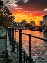 Ein malerischer Sonnenuntergang über einem Fluss in einer Stadt. Die Wolken sind in warmen Farben gefärbt, während die Wasseroberfläche das Licht reflektiert. Am Ufer stehen Bäume und ein Geländer, und im Hintergrund sind Gebäude sichtbar. (Text KI generiert)