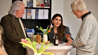Drei Personen stehen an einem Stehtisch. Eine Frau in einem grauen Blazer spricht mit zwei Männern, während sie gestikuliert. Auf dem Tisch stehen eine Flasche Bier und eine Vase mit Tulpen. Im Hintergrund sind Bücherregale sichtbar. (Text KI generiert)