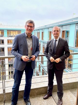 Zwei Männer stehen auf einem Balkon vor einem modernen Gebäude. Der Mann links trägt ein graues Jackett und eine hellblaue Hemd, der Mann rechts ist in einem dunklen Anzug gekleidet. Beide lächeln und halten ihre Hände vor sich. (Text KI generiert)