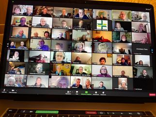 Eine Bildschirmansicht einer Videokonferenz mit vielen Teilnehmern. Die Teilnehmer sind in kleinen Fenstern angeordnet, einige lächeln, andere schauen in die Kamera. Es gibt eine Mischung aus älteren und jüngeren Personen. Einige Fenster zeigen persönliche Hintergründe. (Text KI generiert)