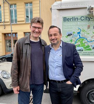 Zwei Männer stehen lächelnd nebeneinander auf einer Straße. Der Mann links trägt eine dunkle Jacke und ein T-Shirt, der Mann rechts einen Anzug mit Hemd. Im Hintergrund ist ein Fahrzeug und ein Schild mit Informationen über Berlin zu sehen. (Text KI generiert)
