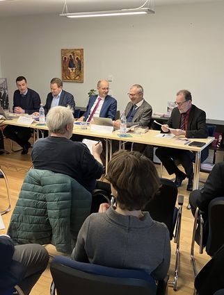 Eine Gruppe von fünf Personen sitzt an einem Tisch in einem Raum. Sie diskutieren, während Zuschauer im Hintergrund sitzen. An der Wand hängen Bilder, und ein Bildschirm zeigt möglicherweise eine Präsentation. Der Raum wirkt informell und ist gut beleuchtet. (Text KI generiert)