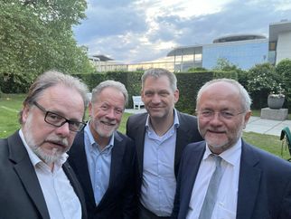 Vier Männer in Anzügen stehen im Freien, umgeben von Grün und einem modernen Gebäude im Hintergrund. Sie lächeln in die Kamera und posieren für ein Gruppenfoto. Der Himmel ist bewölkt, und es ist Abendlicht zu sehen. (Text KI generiert)