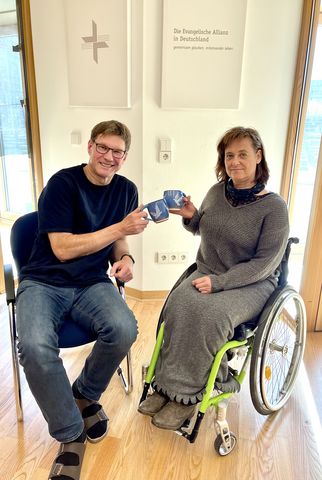 Zwei Personen sitzen in einem Raum. Eine Person im Rollstuhl hält eine Tasse, während die andere Person neben ihr sitzt und ebenfalls eine Tasse hält. Im Hintergrund sind Wände mit Texten und einem Kreuz sichtbar. (Text KI generiert)