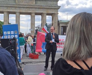 Eine Menschenmenge versammelt sich am Brandenburger Tor. Im Vordergrund steht ein Mann in Anzug und Brille, der spricht. Im Hintergrund sind Banner und Flaggen zu sehen. Einige Personen sitzen auf dem Boden, während andere zuhören. (Text KI generiert)