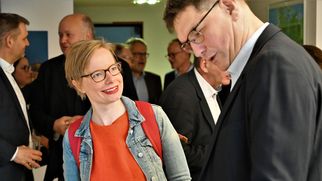 Eine lächelnde Frau mit Brille und einem Jeansjacke steht in einem Gespräch mit einem Mann in Anzug. Im Hintergrund sind weitere Personen in einem informellen Umfeld zu sehen. Die Atmosphäre wirkt freundlich und gesellig. (Text KI generiert)