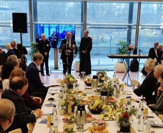 Ein festlich gedeckter Tisch mit verschiedenen Speisen und Getränken. Im Hintergrund stehen zwei Personen, die sprechen, während die Gäste am Tisch aufmerksam zuhören. Die Atmosphäre ist elegant und geschäftlich. (Text KI generiert)