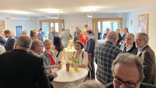 Eine Gruppe von Menschen steht in einem hellen Raum und unterhält sich. Einige Personen stehen an Stehtischen, während andere in kleinen Gruppen zusammenstehen. Es gibt Blumen auf den Tischen und eine entspannte Atmosphäre. (Text KI generiert)