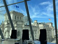 Blick aus einem modernen Konferenzraum auf das Reichstagsgebäude in Berlin. Im Hintergrund sind die Flaggen der Europäischen Union und Deutschlands zu sehen, sowie ein klarer blauer Himmel. Der Raum ist mit einem Tisch und Stühlen ausgestattet. (Text KI generiert)