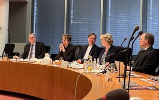 Eine Gruppe von fünf Personen sitzt an einem runden Tisch in einem Konferenzraum. Sie diskutieren, während Wasser und andere Getränke auf dem Tisch stehen. Im Hintergrund sind große Fenster mit Jalousien zu sehen. (Text KI generiert)