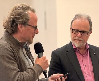 Zwei Männer stehen auf einer Bühne. Der Mann links spricht ins Mikrofon und hält ein Smartphone in der Hand, während der Mann rechts ihn aufmerksam ansieht. Beide tragen formelle Kleidung und scheinen an einem Gespräch oder einer Diskussion beteiligt zu sein. (Text KI generiert)