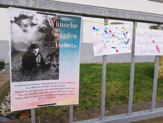 Ein Plakat mit der Aufschrift "Wünsche werden Gebete" und einem Bild einer betenden Person. Daneben hängt ein buntes Papier mit handschriftlichen Wünschen und bunten Fingerabdrücken. Im Hintergrund ist ein grüner Rasen zu sehen. (Text KI generiert)