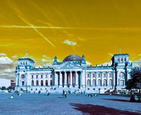 Das Bild zeigt das Reichstagsgebäude in Berlin, das durch eine gelb-orangefarbene Filterung hervorgehoben wird. Im Vordergrund sind Menschen auf einer Wiese zu sehen, die das Gebäude umgeben. Der Himmel ist teilweise bewölkt mit sichtbaren Flugzeugstreifen. (Text KI generiert)