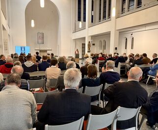 Eine Gruppe von Menschen sitzt in einem hellen, modernen Raum mit hohen Decken. Im Vordergrund steht eine Rednerin, während die Zuhörer aufmerksam in ihre Richtung schauen. Die Atmosphäre wirkt konzentriert und respektvoll. (Text KI generiert)