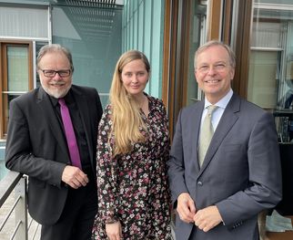 Drei Personen stehen auf einem Balkon vor einem modernen Gebäude. Sie lächeln und posieren für das Foto. Die Frau in der Mitte trägt ein blumiges Kleid, während die beiden Männer formelle Kleidung tragen. Im Hintergrund sind Fenster und eine Überdachung sichtbar. (Text KI generiert)