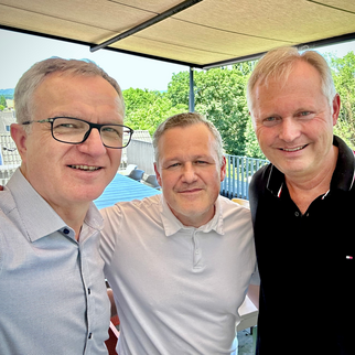 Drei Männer stehen lächelnd zusammen auf einer Terrasse. Sie tragen unterschiedliche Hemden und stehen vor einer grünen Landschaft. Die Atmosphäre wirkt freundlich und entspannt.