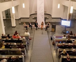 Eine Veranstaltung in einem großen Raum mit Sitzreihen. Auf der Bühne sitzen fünf Personen, die an einem Podiumsgespräch teilnehmen. Im Hintergrund ist ein Bildschirm sichtbar. Das Publikum sitzt aufmerksam und schaut in Richtung Bühne. (Text KI generiert)
